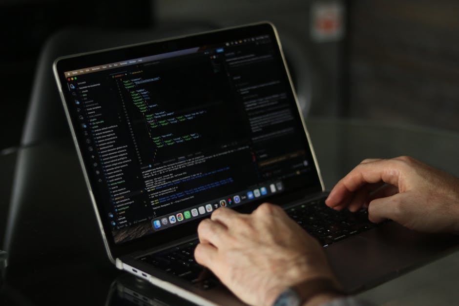 A close-up of a person's hands typing on a laptop computer, with the screen displaying a dark-themed coding interface filled with lines of programming code in various colors. The laptop is placed on a